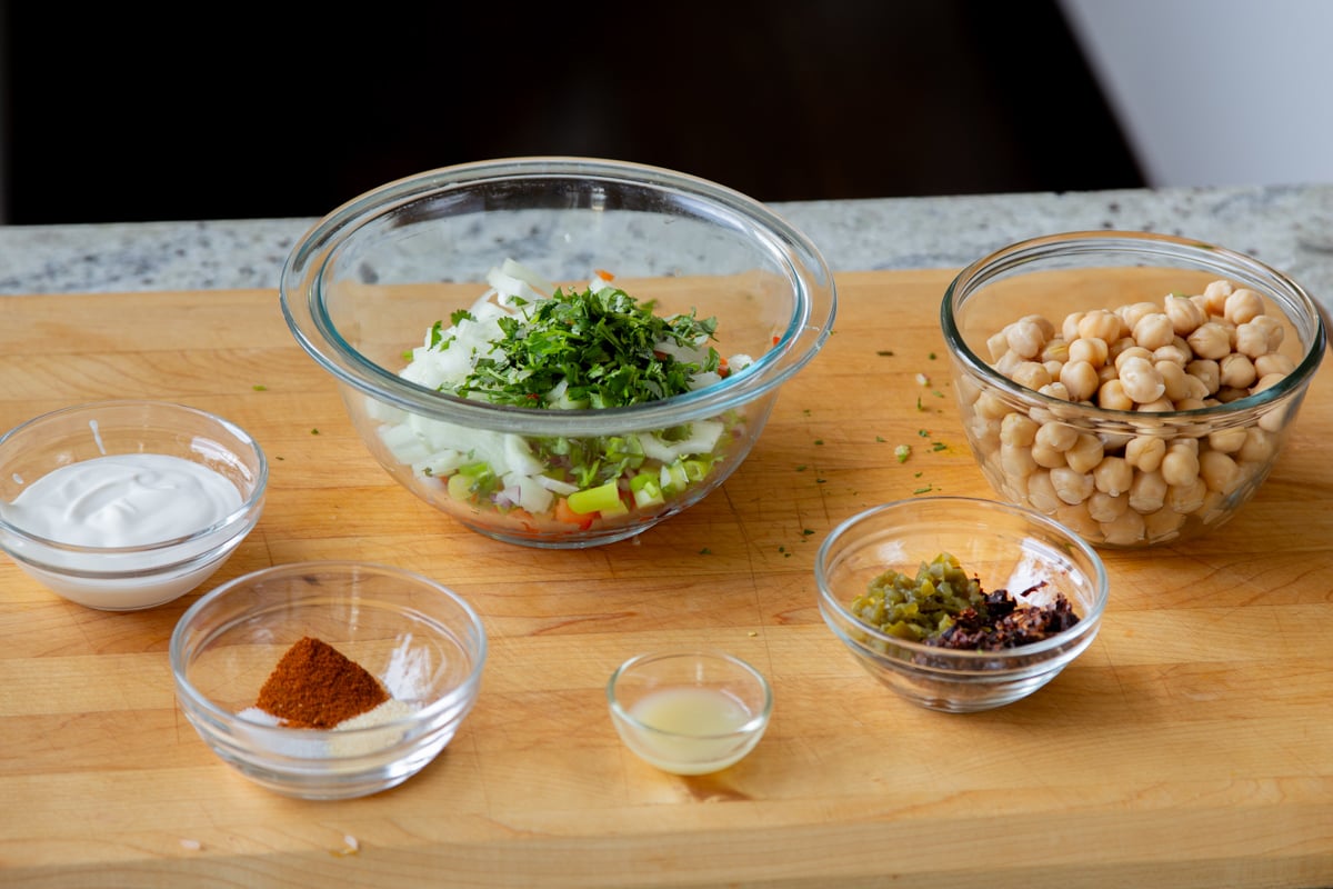chickpeas, veggies, and other salad ingredients in bowls on a wooden cutting board