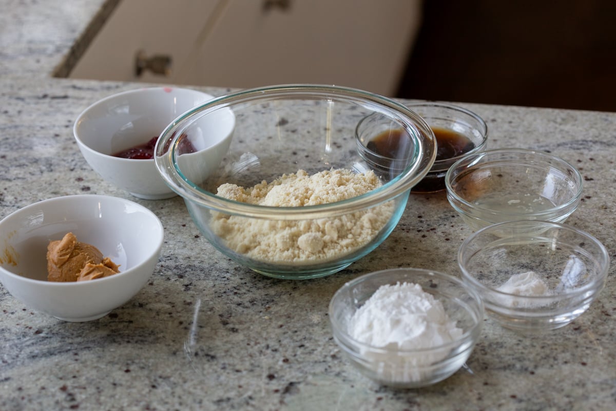 almond flour, peanut butter, jam, and other cookie ingredients in bowls on a kitchen counter