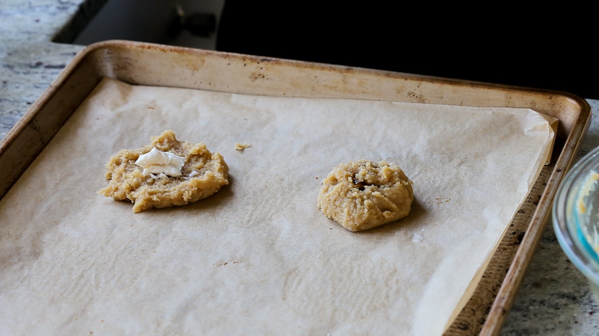 2 unbaked cookies on a baking sheet, one closed up and one with a dollop of cream cheese mixture on it