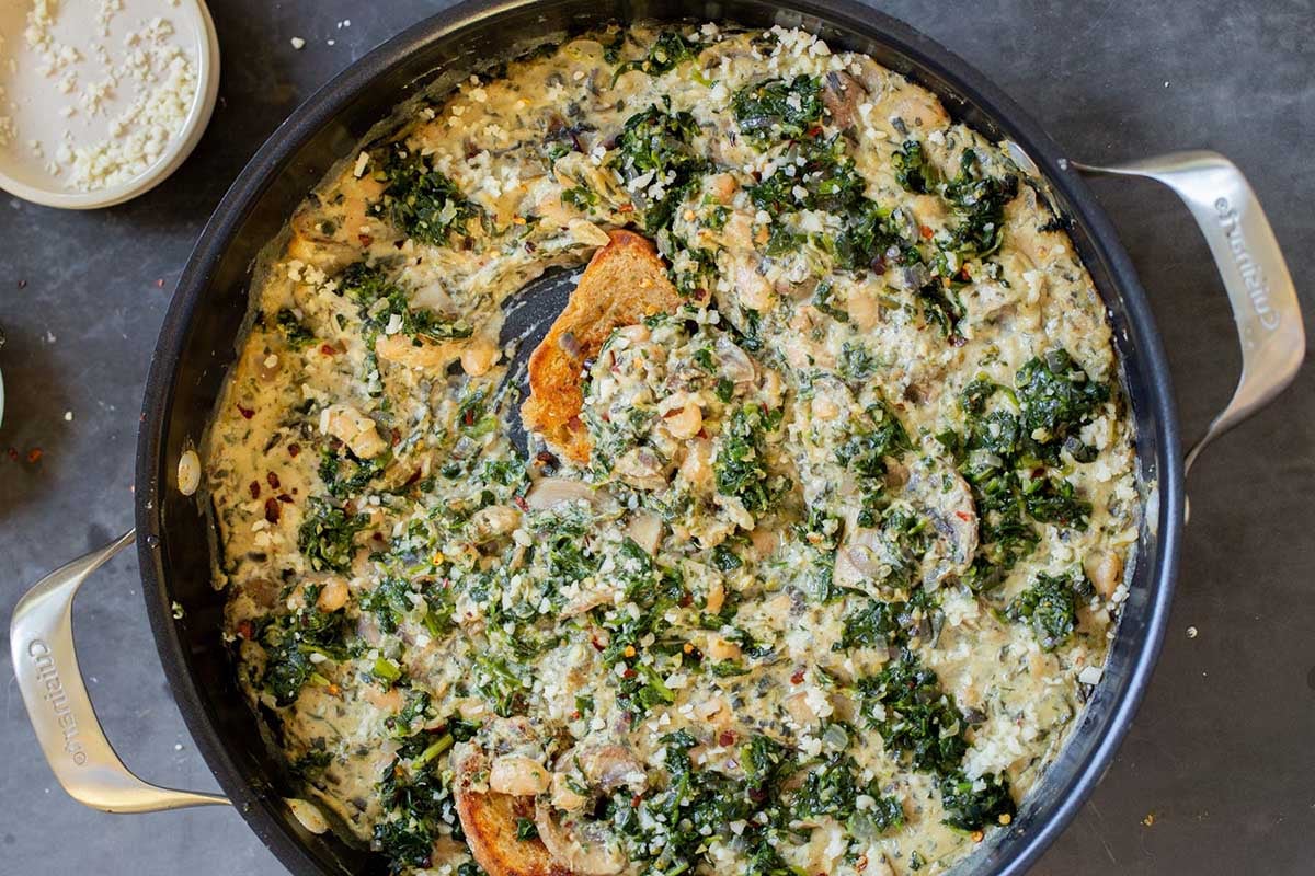 dipping a piece of bread into vegan white bean mushroom alfredo in the pan with spinach
