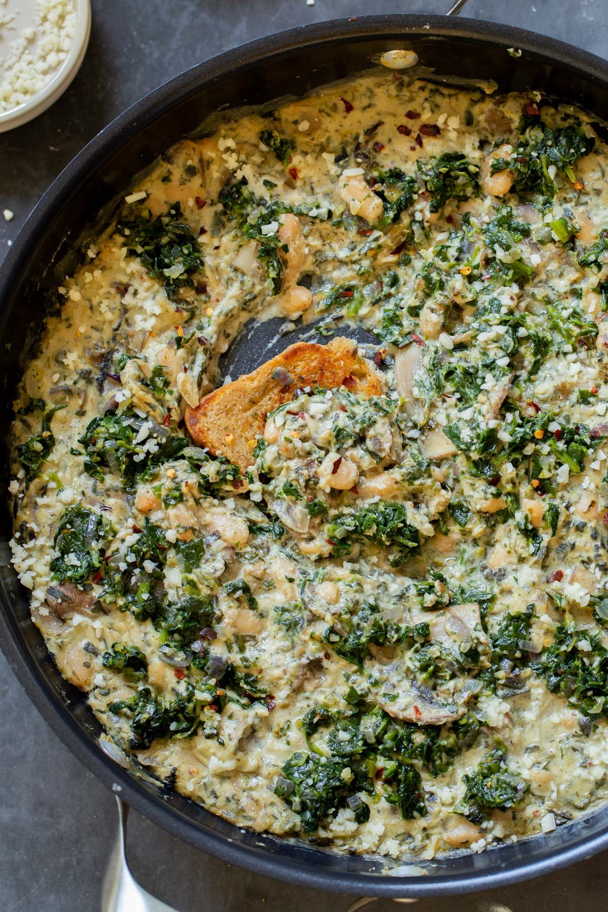 dipping a piece of bread into vegan white bean mushroom alfredo in the pan with spinach
