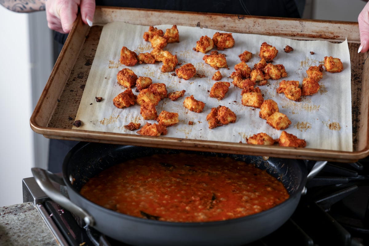 tofu coated in cornstarch and spices on the baking sheet after baking, held over the pan of madurai chicken curry sauce