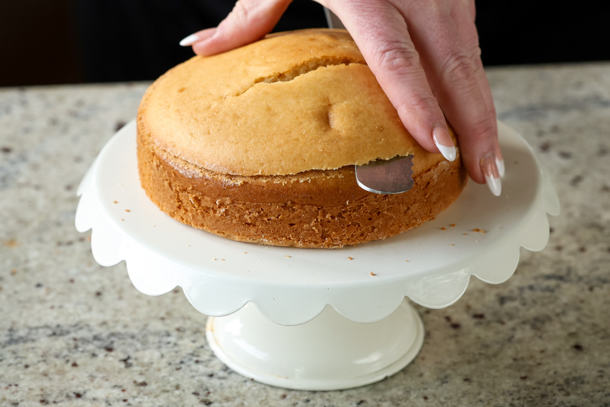 slicing the top off of the cake on a cake stand after baking