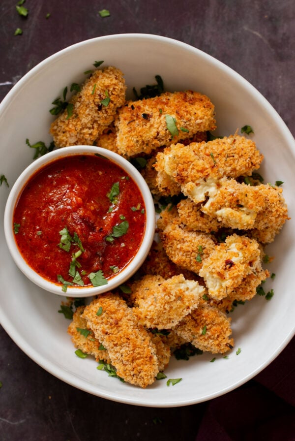 vegan mozzarella sticks in a serving platter with marinara on the side with some broken sticks, so you can see the cheese