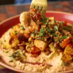 fork taking a bite of harissa bean puree topped with roasted vegetables and baharat walnuts